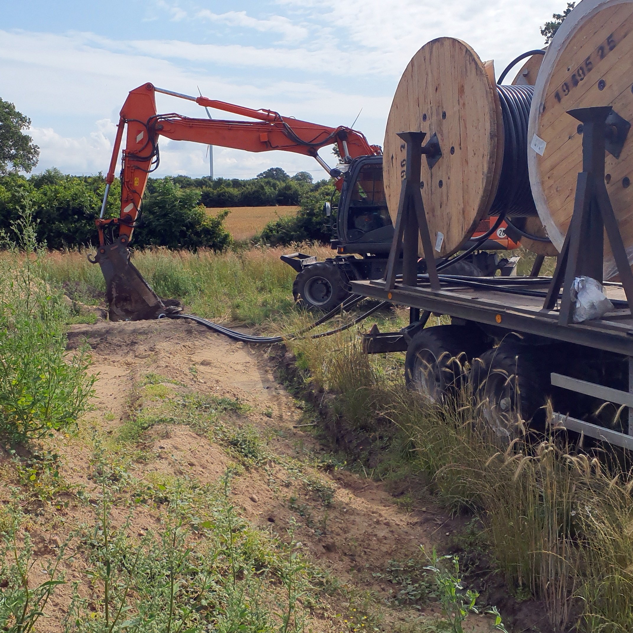 An excavator is standing behind a trailer loaded with cable reels on which power cables are wound. In the background are bushes, trees and a field.