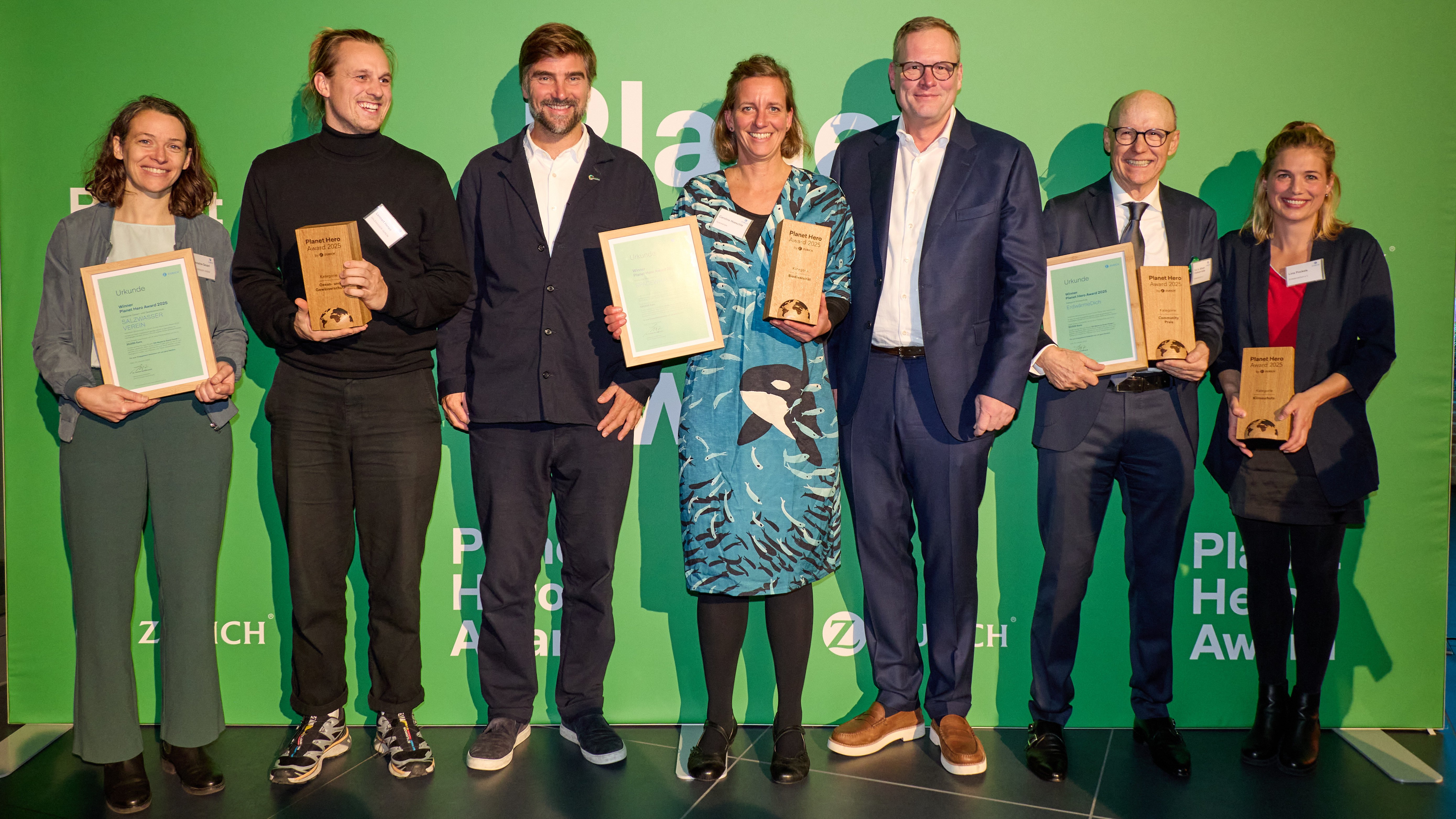 Seven adults stand in front of a green background and smile at the camera. Some of them are holding award certificates in their hands.