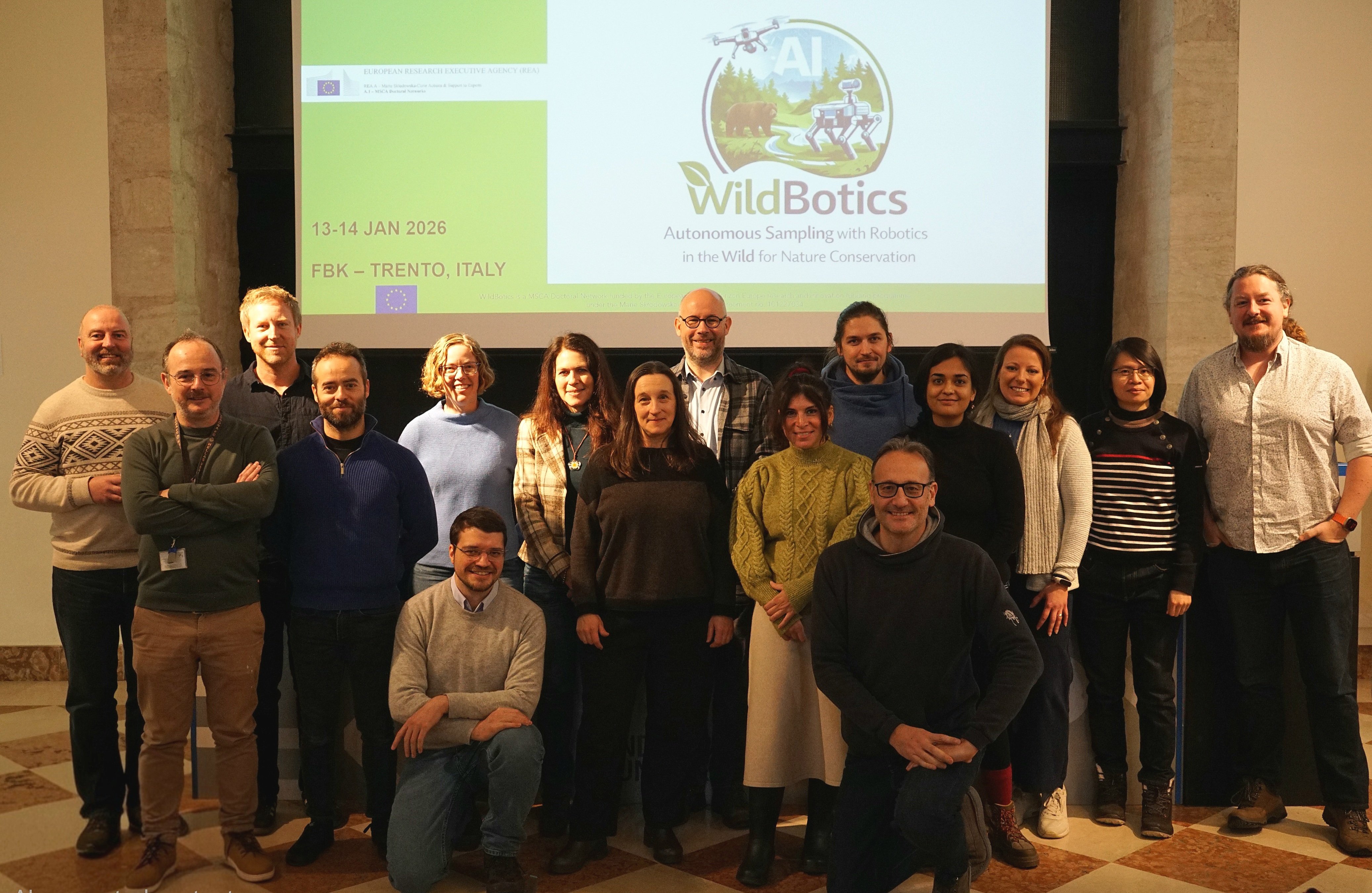 17 Menschen stehen für ein Gruppenfoto vor einer Leinwand, auf der unter anderem das Wort "WildBotics" zu lesen ist.