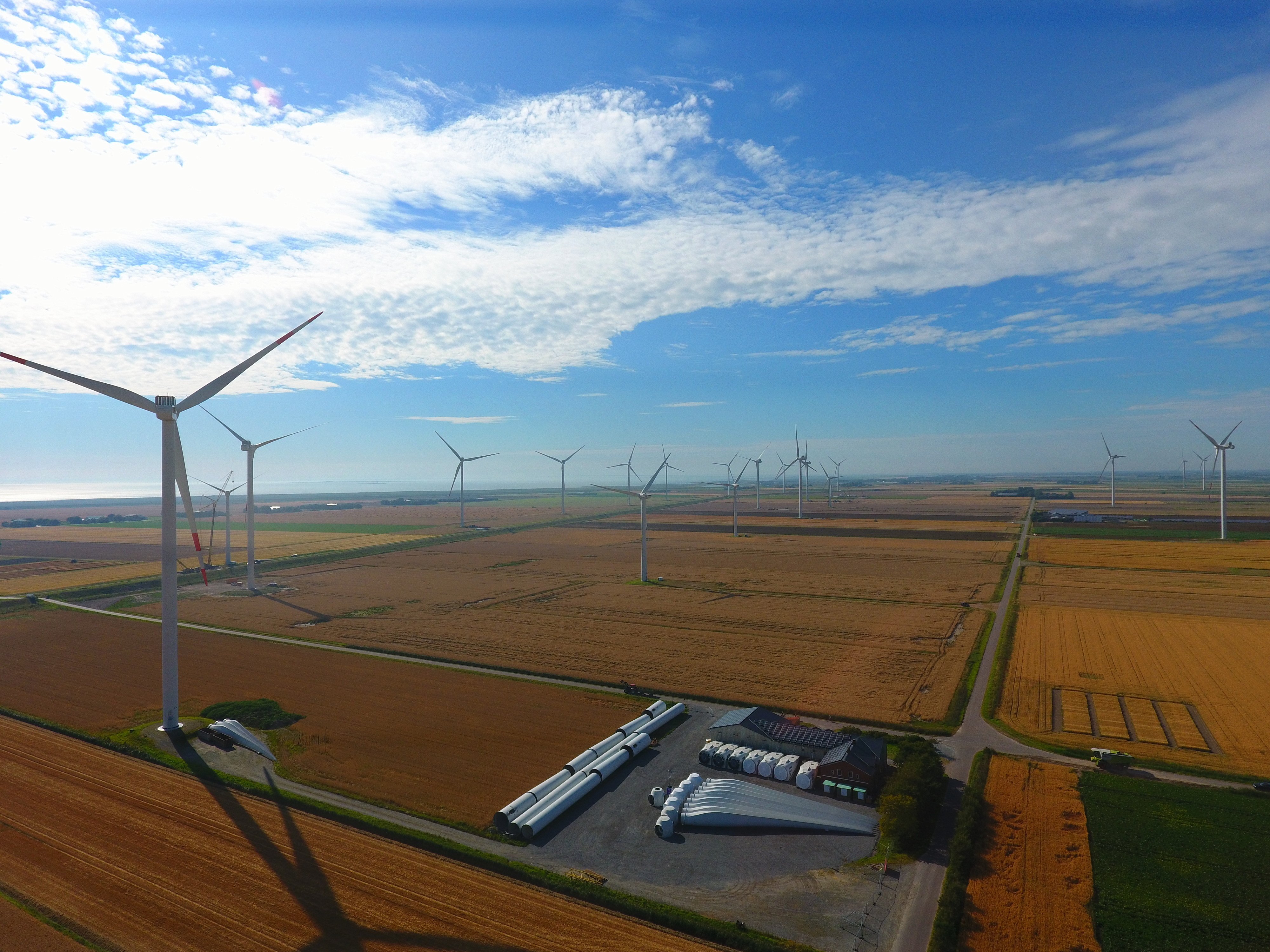 Luftaufnahme über Felder, auf denen Windräder stehen. Im Vordergrund werden Bauteile für Windenergieanlagen gelagert.