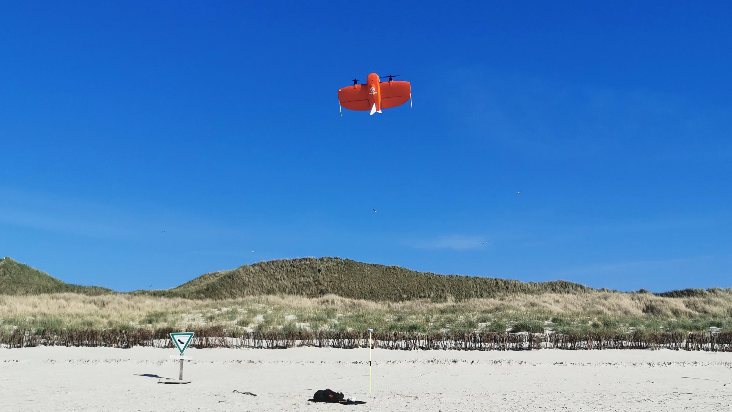A drone takes off against a blue sky on a beach. Dunes can be seen in the background.
