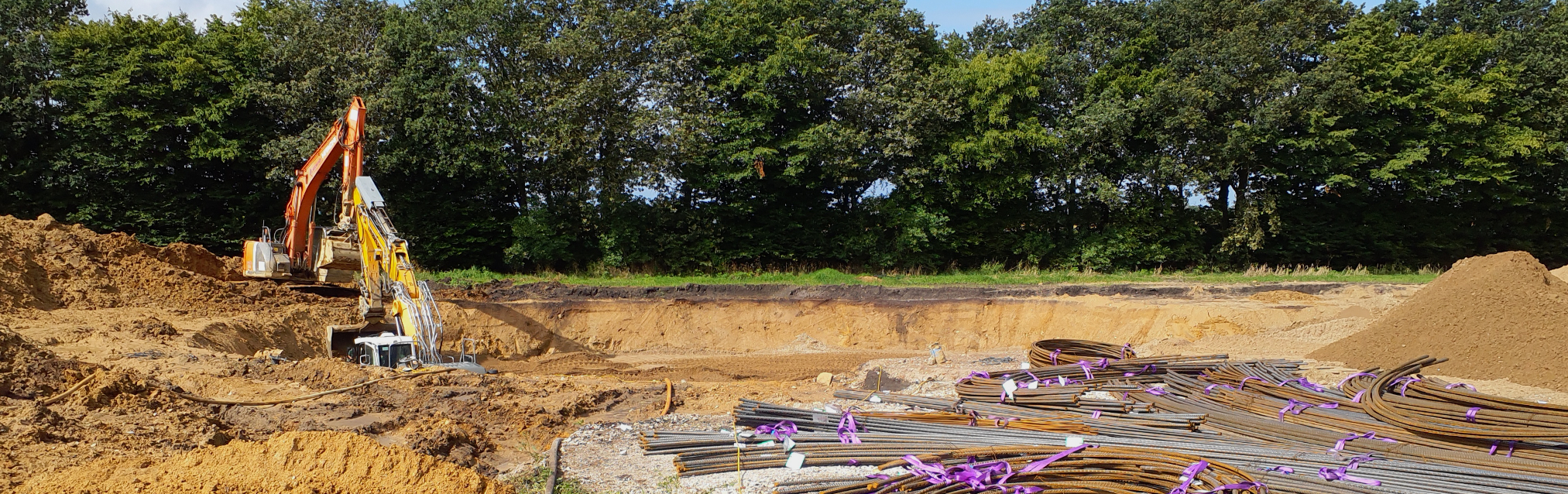 A construction site with two excavators in a large excavation pit. Reinforcing steel bars lie in the foreground, whilst trees stand in the background.